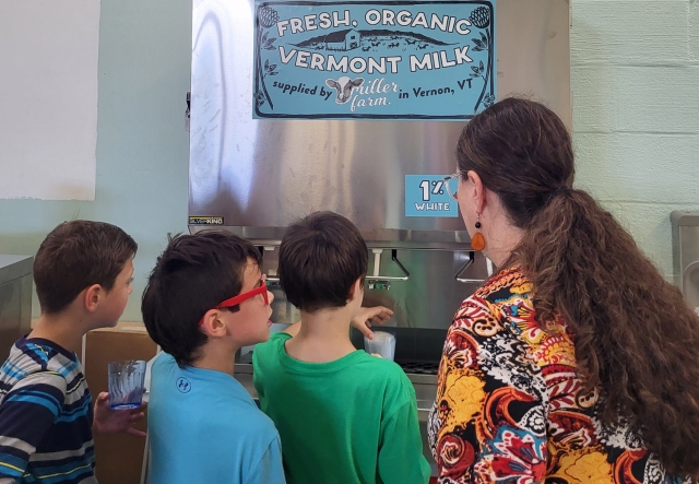 Three children and one adult at a milk dispensing station with a sign reading "Fresh, Organic Vermont Milk supplied by Miller Farm in Vernon, VT."