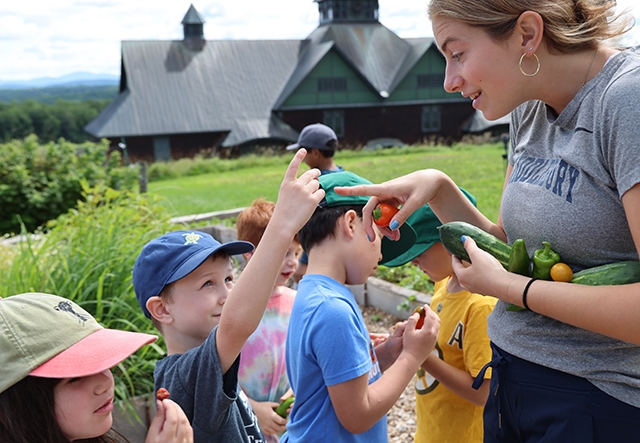 A group of children gather around an educator in an outdoor garden holding freshly harvested vegetables and fruit