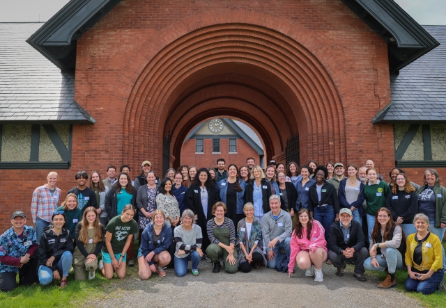 Sixty adults and young adults smile while wearing nametags posed in rows in front of a large brick barn