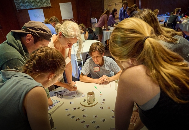 Six adults stand and sit around a circular table, looking down at small cards spread out, during a professional learning exercise