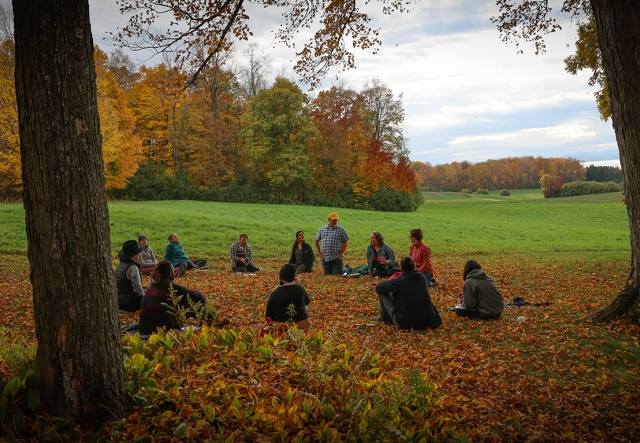 A circle of twelve people sit on a leaf-covered lawn in autumn with sweeping views of autumn foliage and rolling pastures in distance