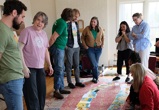 A group of adults in discussion in a living room gathered around pages of a picture book spread out on the floor
