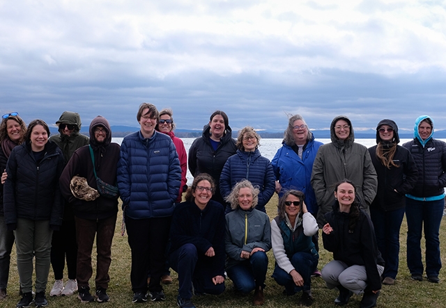 A group of 15 adults pose for a photo in front of a lake