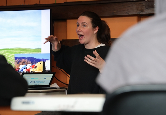 A young woman teaches animatedly in a classroom in front of a screen