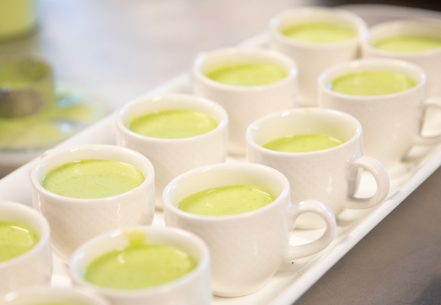 A tray of white cups filled with green soup, arranged in a neat row on a counter.