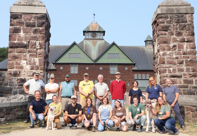 17 people in two rows: one group kneeling, one standing behind them. There's a dog and a calf in the front row. Everyone is standing in the gravel entrance between two stone pillars of the Farm barn