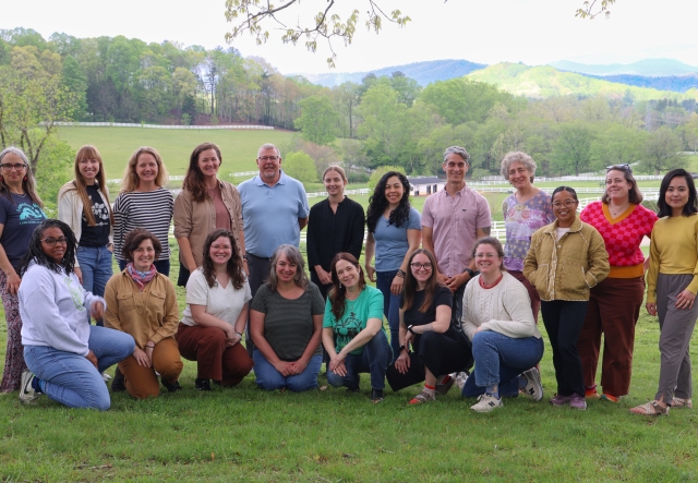 A large group of adults pose for a photo, a scenic farm landscape in the background.
