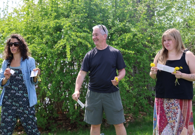 Three adults stand outdoors in spring holding flowers and index cards during an activity