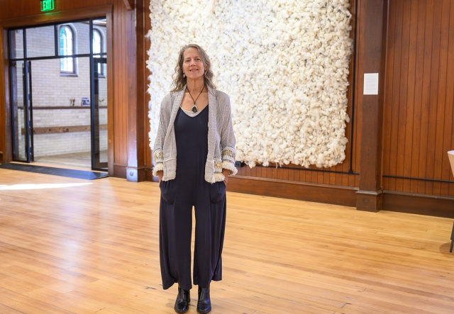 woman in gray sweater and black pantsuit stands in front of a large hanging of woven wool.