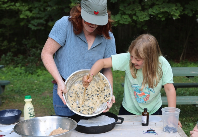 girl scrapes blueberry cake dough from a bowl held by an adult, into a cast iron pan.