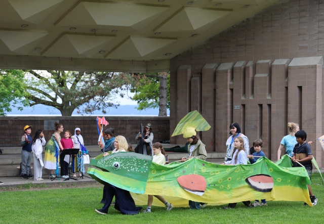 Young children parade in a large trout puppet across an outdoor amphitheater