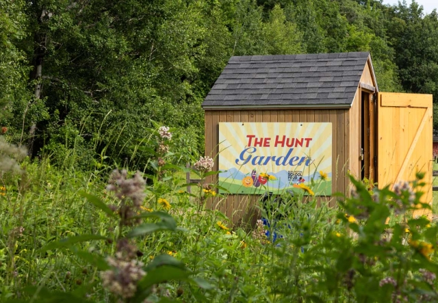 Small wooden shed featuring a sign that reads "The Hunt Garden," surrounded by lush greenery and wildflowers. 