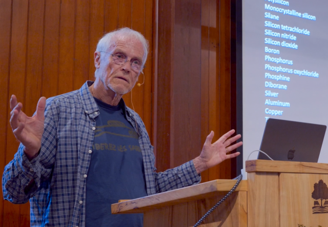 white-haired man in glasses, with open, button-up blue plaid shirt over blue shirt, stands at a podium talking with arms open.