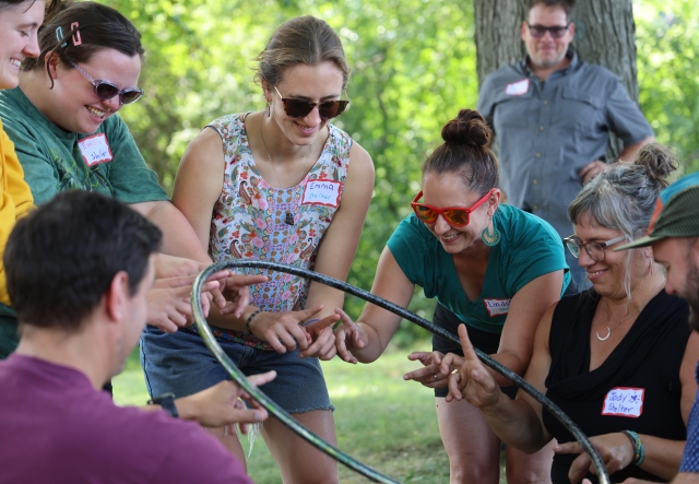 Seven people smile while balancing a hula hoop on their hands in an outdoor team building activity