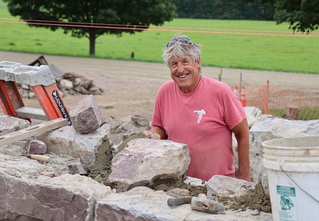 man with salt and pepper hair holding masonry trowel stands behind a half-built stone wall