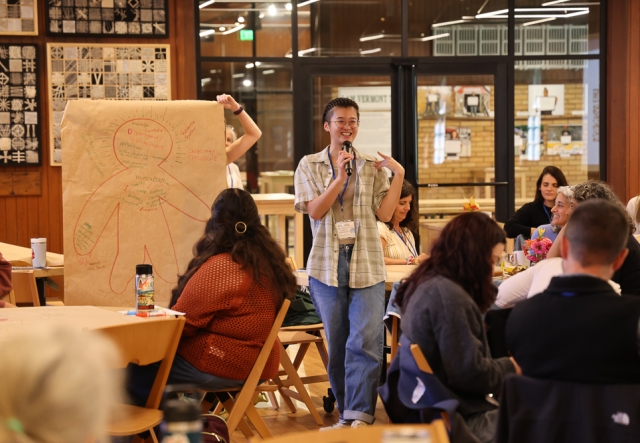 A person smiles while talking into a microphone in a large hall at Shelburne Farms Coach Barn