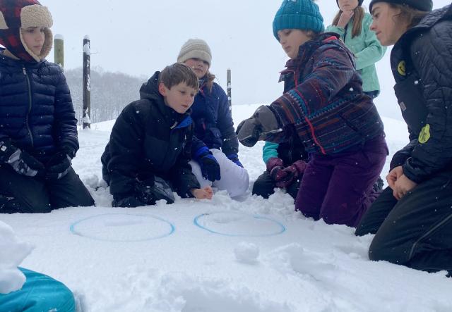 Four students kneel on snowy ground in winter inspecting animal tracks