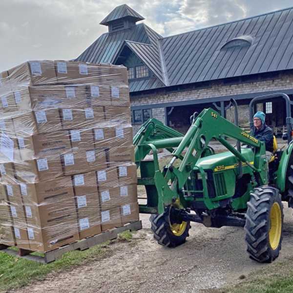 man in green tractor lifting stack of shrink-wrapped cardboard cartons.