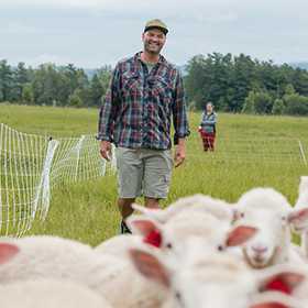 Educator Jed Norris walks in a grassy field behind a flock of sheep