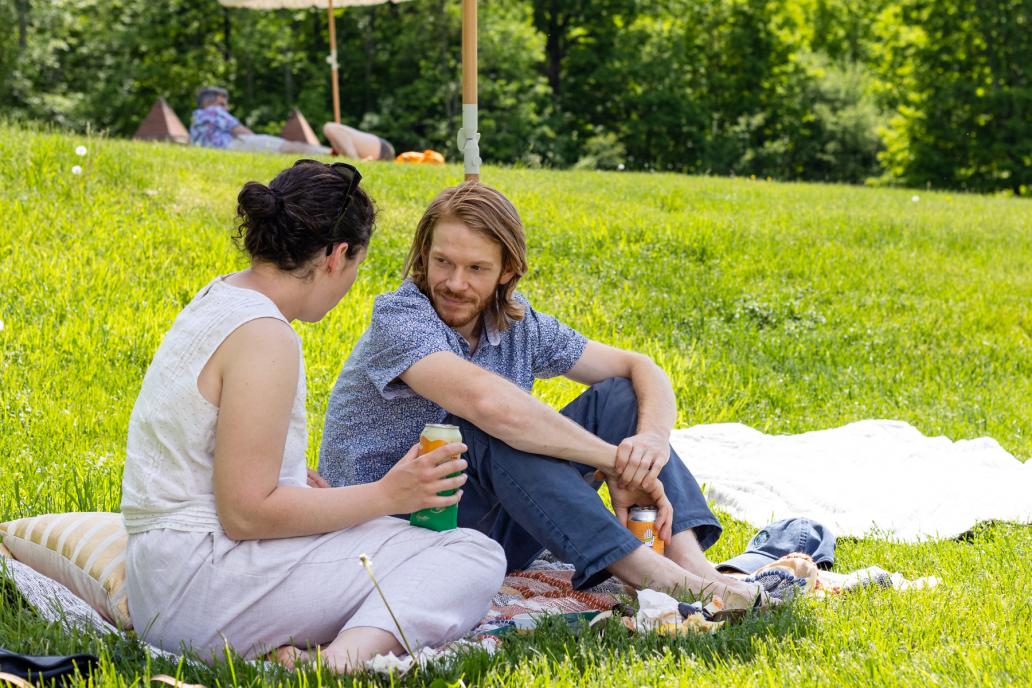Two people sit under a large umbrella on a lawn, enjoying a picnic.