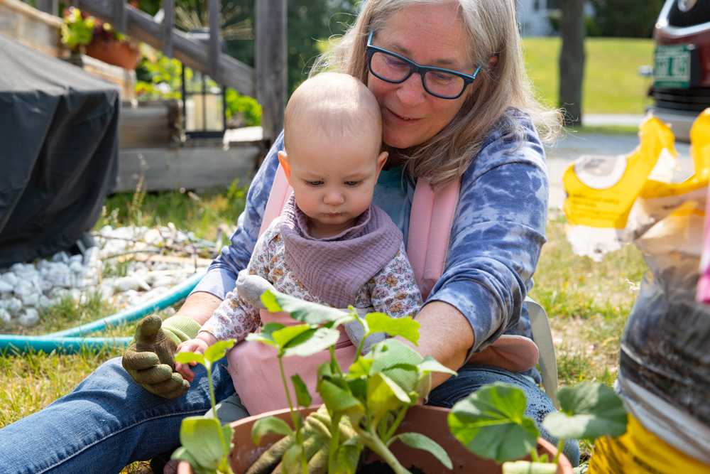A woman sits with a baby in a carrier attached to her body, they look at a squash plant