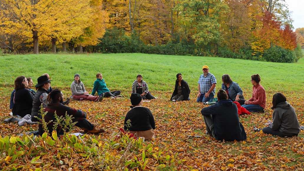 Sixteen adults sit in a circle outside on a grassy lawn in discussion with fall foliage in the background