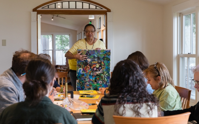 A half dozen people sit around a dining room table in a cozy room with white walls, with a woman standing at the head of the table, holding a collaged piece of artwork