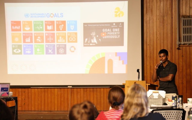 A young person holds a microphone at a podium speaking to a large ballroom with Sustainable Development Goals on a screen on far wall