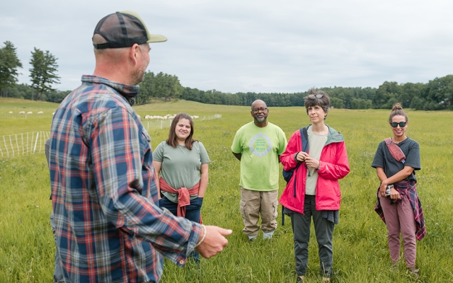man in cap and flannel shirt facing away from camera talking to four people (long pants, short sleeves), all standing in a grass pasture.  