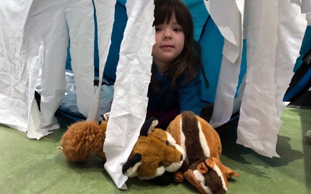 Child peeks out of a fort with hanging tissue paper sides with two fox stuffed animal toys.