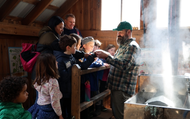 Person in sugarhouse gives a demo to a group of children and adults.