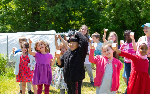 A group of smiling children holding up cups in a sunny outdoor setting, with greenery and a fence in the background.