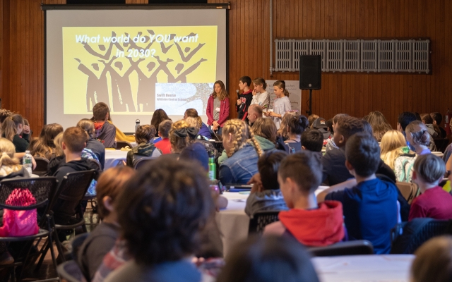 A group of young people stand in front of a projector screen that reads, what world do you want in 2030, and speak to a large room of people