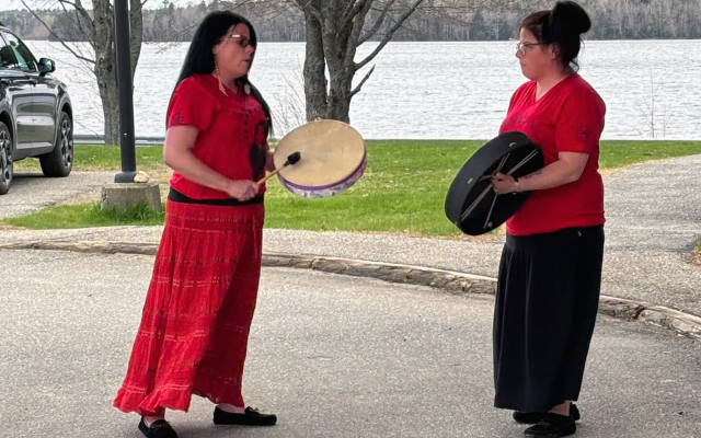 Two women wearing red playing hand drums in front of a body of water.