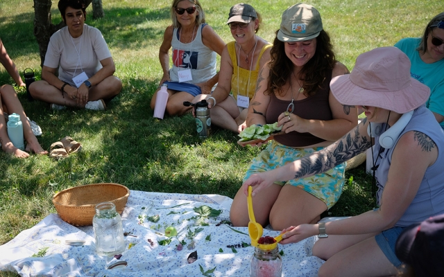 A small group of women smile while seated around a blanket in the grass, making an herbal tea during a workshop