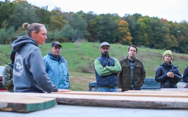 People gather around a collection of lumber outdoors, one person speaks to the group.