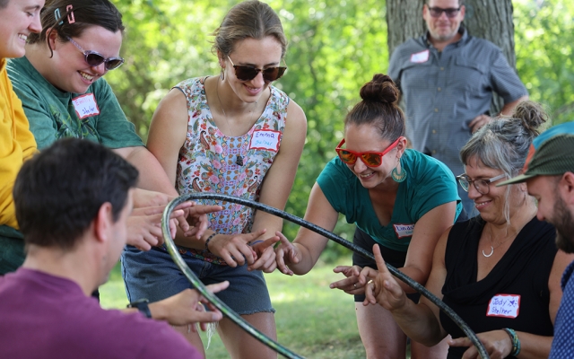 Seven people smile while balancing a hula hoop on their hands during a team building activity