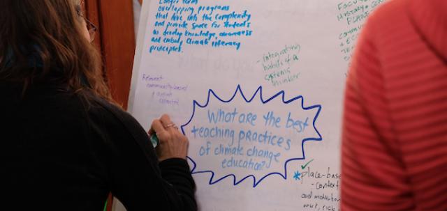 A flipchart hands on an easel, seen over two educators' shoulders. One educator writes a response to a question written on flipchart, what are the best teaching practices of climate change education?