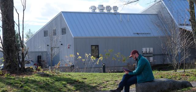 Educator sits alone on a rock outside, writing in a notebook. The Maine Ecology School's gathering center is in background.
