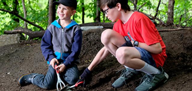 Two young students dig in school garden with trees in background. One student smiles off camera while holding gardening tool.