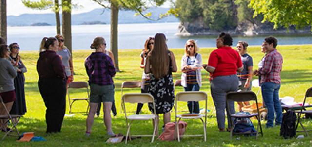 A circle of educators stand in grass with Lake Champlain in background. They engage in a group activity.