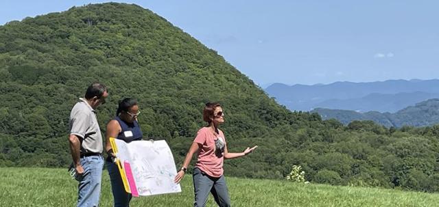 Three educators conduct presentation on mountaintop. One educator holds a posterboard while another gestures while talking. Green mountains stand in background against a blue sky.