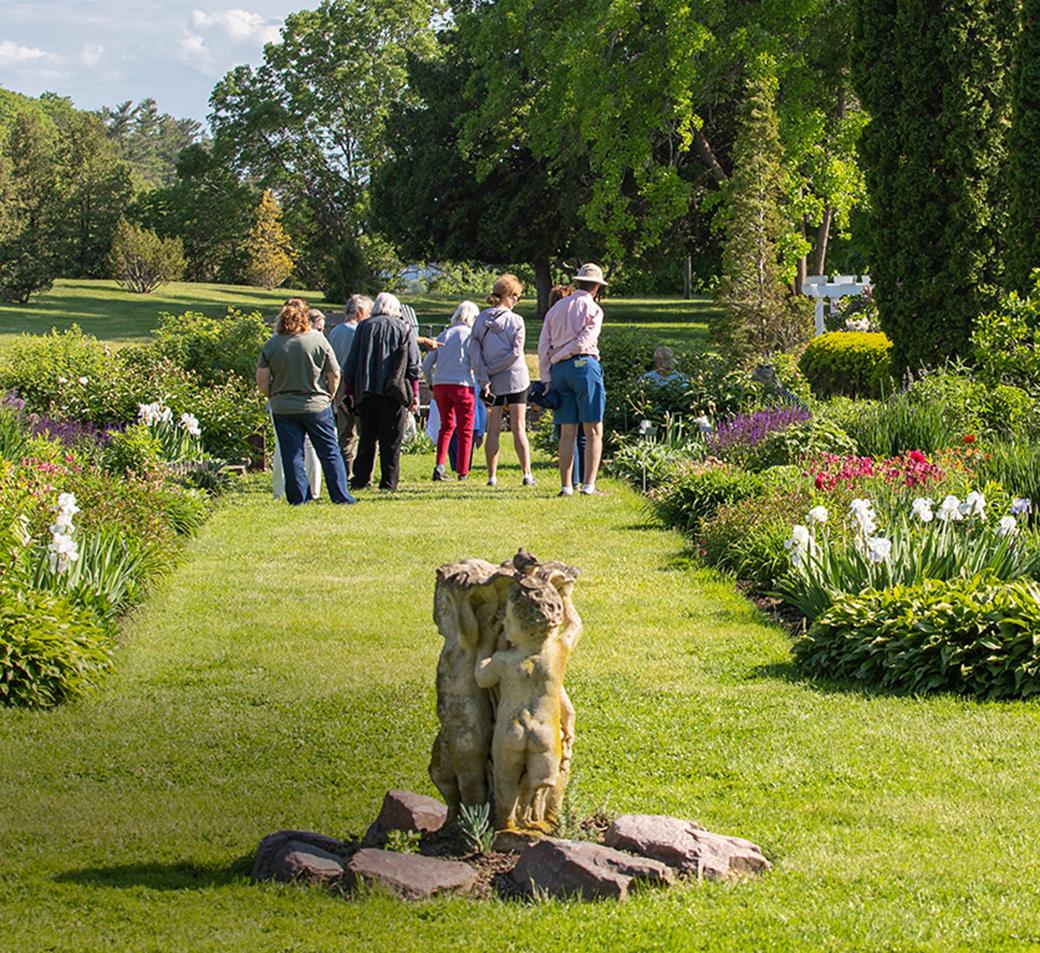 group of people walking down grassy path in flower garden with statue in foreground