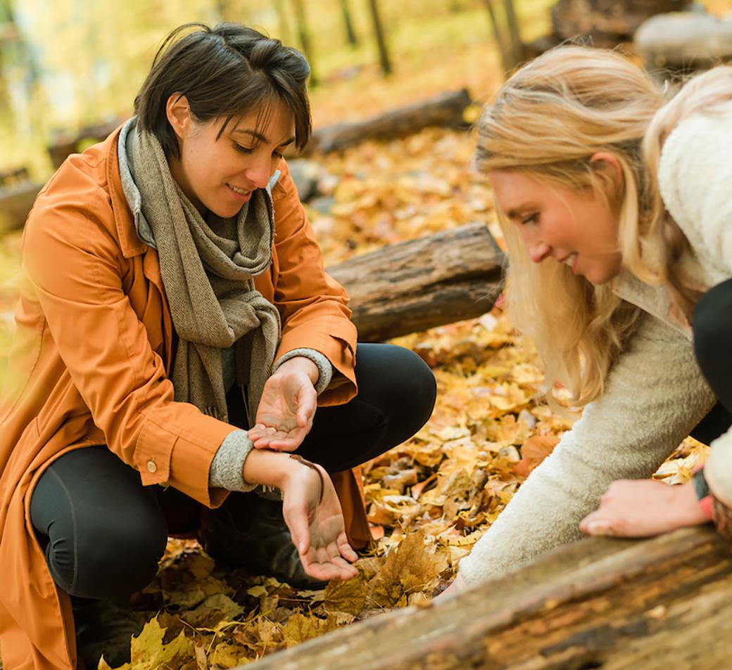 Two educators kneel and smile while carefully handling and admiring a newt on the forest floor at Shelburne Farms in autumn. Logs and leaves cover the ground around them. 