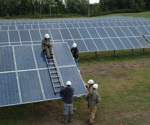 A crew of workers in hard hats install solar panels in front of rows of other solar panels in a green field.