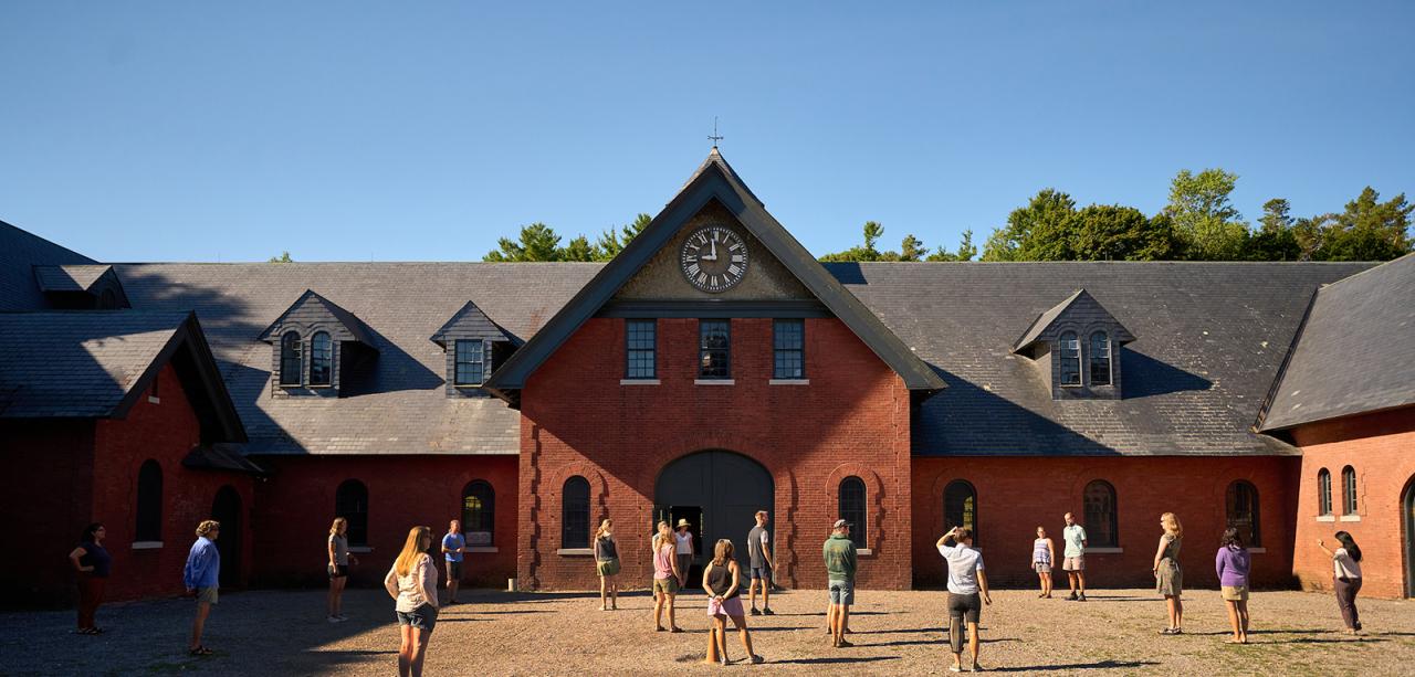 A wide view of a gravel courtyard in summertime surrounded by a historic brick building, the Shelburne Farms Coach Barn. Two dozen educators stand in the courtyard.