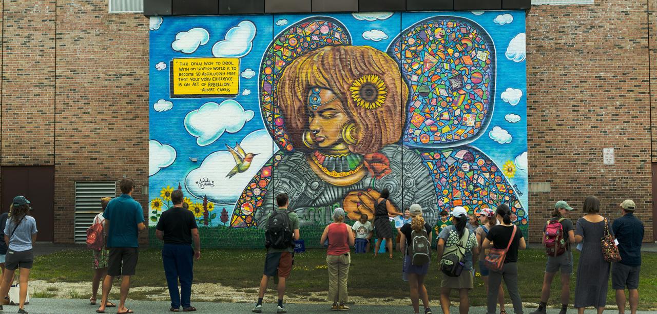 A dozen educators stand with backs to camera. They admire a one-story mural on a brick wall at Champlain Elementary School of Kelis the Afronaut.