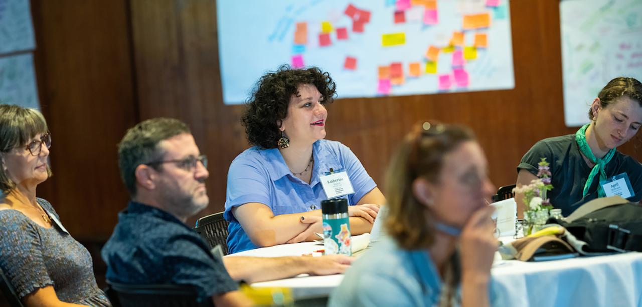 A half dozen educators sit around a small table at Shelburne Farms' Coach Barn, turned to watch a presentation. Educator at center smiles with post-it notes on wall in background.