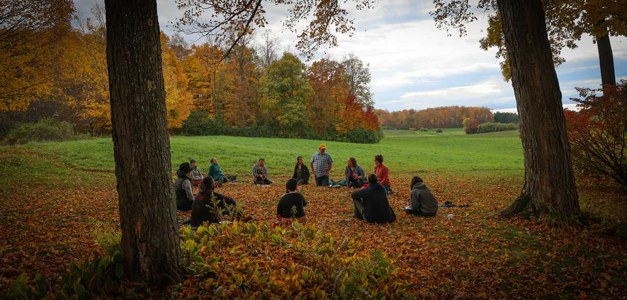 A circle of twelve people sit on a leaf-covered lawn in autumn with sweeping views of autumn foliage and rolling pastures in distance
