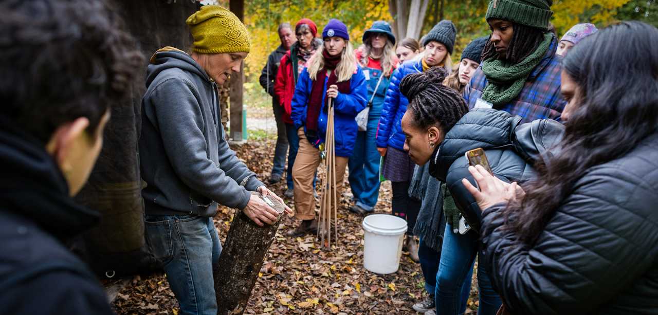 Two dozen educators stand in a circle listening to an instructor, who holds a log growing shiitake mushrooms, outside of a cabin in fall